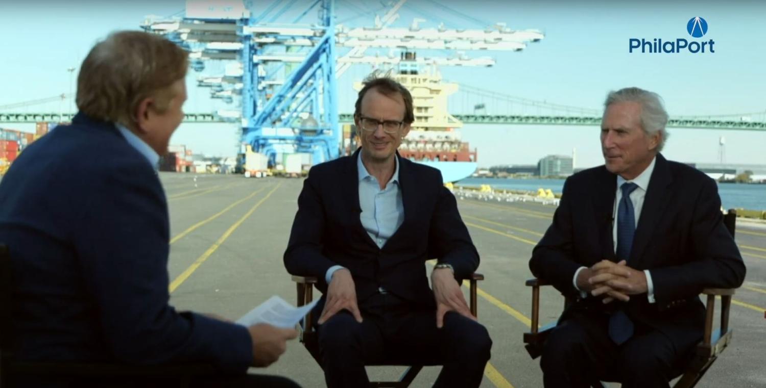 Three men in suits sitting in director's chairs at the Port of Philadelphia with a large crane in the background